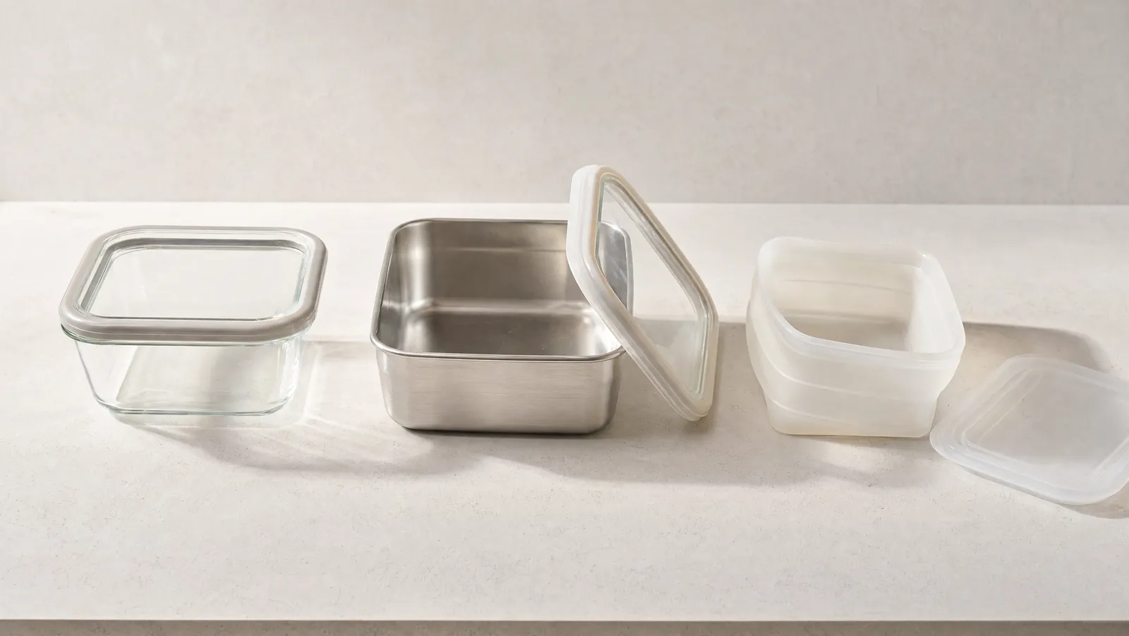Three empty food storage containers in a row on a bone-white counter in soft raking daylight: a square clear glass box with a glass lid in a pale silicone frame resting flat on top, an open brushed stainless steel box with its clear glass lid propped against its side so the bare metal interior shows, and a translucent milky silicone box with the concertina folds readable in its sidewall and its soft lid lying beside it