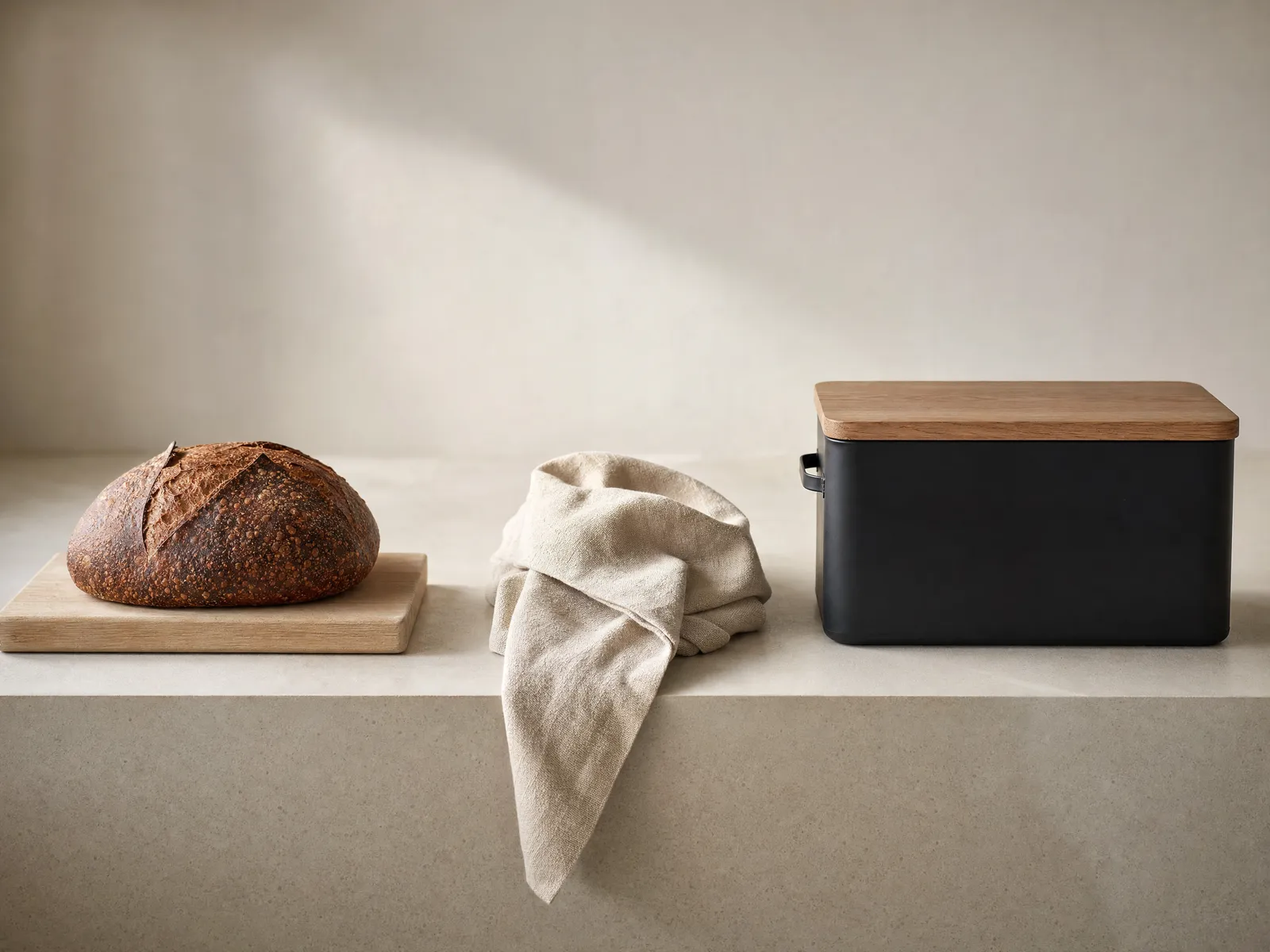Three plastic-free ways to keep a loaf, on a bone-white counter in soft daylight: a round crusty scored sourdough loaf resting on a pale wooden board, a loosely folded undyed linen cloth with one corner draped over the counter edge, and a closed matte black steel bread bin with a plain wooden lid sitting flush on top
